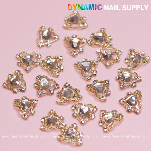A pink surface is adorned with luxury heart-shaped nail charms featuring gold beading and clear rhinestones, ideal for a Valentine nails design. Each charm is surrounded by shiny crystals, adding a delicate and elegant touch. The brand name Dynamic Nail Supply is displayed at the top of the image.