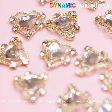 Close-up of the luxurious 20 pcs Luxury Heart Charm for Valentine Nails Design by Dynamic Nail Supply, featuring gold borders and rhinestone accents. The hearts, each adorned with a central faceted gem, are beautifully arranged on a pink surface. In the top right corner, "Dynamic Nail Supply" is displayed in colorful letters, making it perfect for your Valentine nails design. The website URL is placed at the bottom.