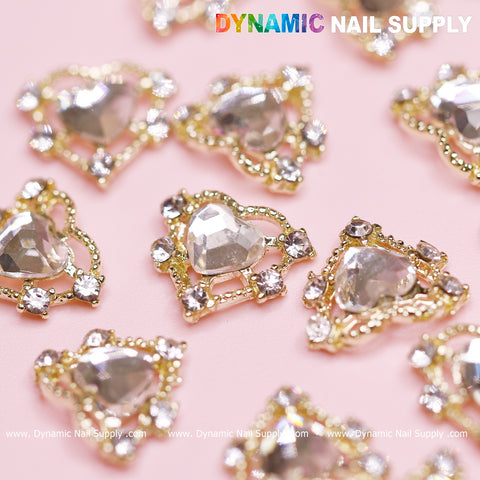 Scattered on a light pink surface are luxury heart-shaped charms, featuring clear, faceted centers surrounded by a rhinestone border with gold detailing. The Dynamic Nail Supply logo is prominently displayed at the top, enhancing the elegance of this Valentine's Day nail design.