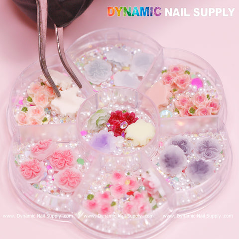 A circular nail supply tray from Dynamic Nail Supply, with six compartments, beautifully organizes a variety of nail art decorations, including small 3D flowers in shades of pink and white and tiny iridescent beads. A pair of tweezers hovers over the 40-piece set featuring White and Pink 3D and 5D Rose Flower Charms with pearls, ideal for Valentine nails art design, as they select a white floral piece against a light pink background.