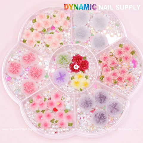 A circular tray with multiple compartments displays a charming Valentine's nail art design, featuring various 3D floral decorations in pink, red, yellow, purple, and gray shades. Highlighted with small pearls and rhinestones, this creation is crafted using the "40 pcs White and Pink 3D 5D Rose Flower Charm with Pearls Accessories for Valentine Nails Art Design" by Dynamic Nail Supply.