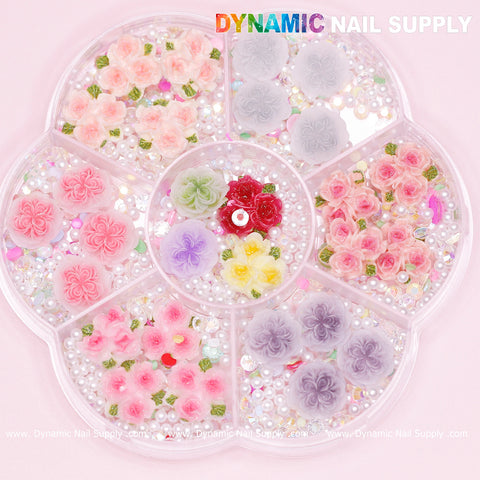 A circular flower-shaped container from Dynamic Nail Supply holds a variety of decorative nail art pieces. Each section showcases Valentine Nails Art Designs featuring colored rose designs, including pink and white 3D and 5D roses, complemented by small pearls and beads. The container is labeled with the brand name on a pastel pink background.