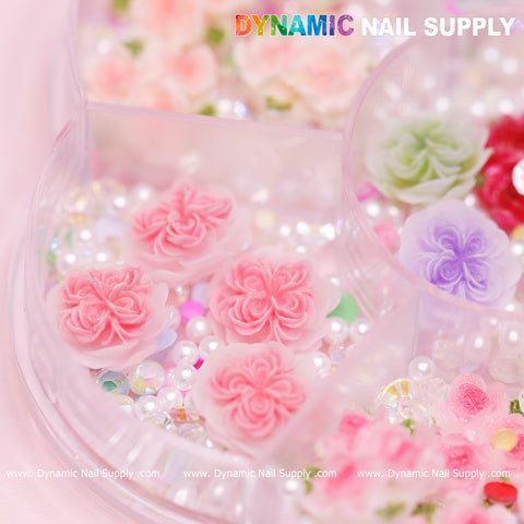A close-up of the 40 pcs White and Pink 3D 5D Rose Flower Charm with pearls from Dynamic Nail Supply showcases pastel-colored flower embellishments, including pink and purple blossoms, ideal for Valentine nails art design. Nestled among various sizes of small iridescent beads, you'll find charming 3D rose flowers in a neatly sectioned container.