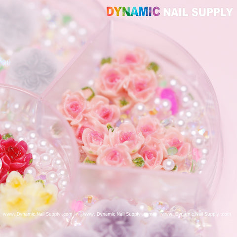 A detailed view of the Dynamic Nail Supply product highlights a container filled with 40 pieces of pink and white 3D and 5D rose flower charms, complete with pearl accessories, ideal for creating Valentine-themed nail art. The container features separate compartments and the brand’s name, Dynamic Nail Supply, displayed in lively text against a soft pink background.