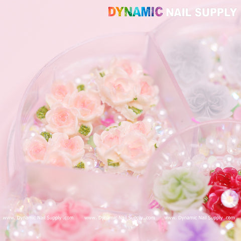 A close-up of a nail art accessory container showcases compartments filled with the Dynamic Nail Supply's 40 pcs White and Pink 3D 5D Rose Flower Charm with pearls, perfect for Valentine nails art design. The roses, accompanied by green leaves and tiny white pearls, are beautifully displayed in the partially transparent container branded with Dynamic Nail Supply.
