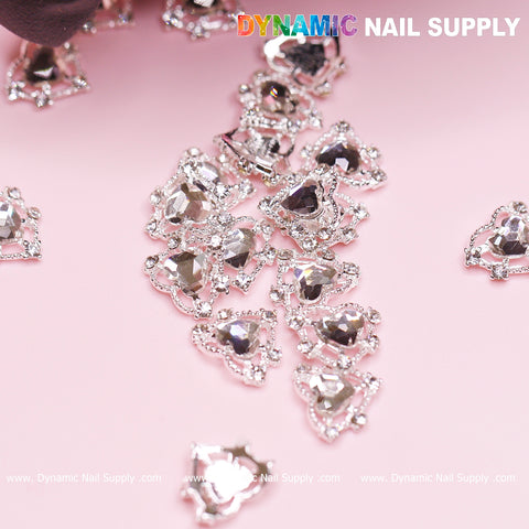 A set of 20 exquisitely detailed silver heart charms, featuring crystal rhinestones ideal for a Valentine Nails Design, displayed on a soft pink background. These charms are adorned with sparkling, faceted surfaces surrounded by tiny clear gems. The Dynamic Nail Supply brand name is prominently displayed at the top, complemented by a watermark below.