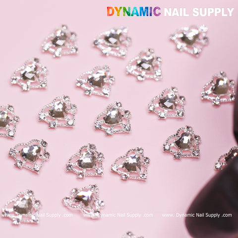 A detailed view shows a collection of silver heart-shaped rhinestone charms neatly arranged on a pink surface. Designed specifically for Valentine Nails Design, each charm highlights a prominent central gem encircled by smaller stones. The branding, "Dynamic Nail Supply," is prominently displayed along the image's edges.