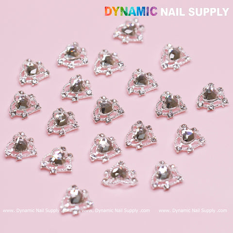 Close-up of several silver heart-shaped rhinestone nail decorations arranged on a light pink surface, perfect for a Valentine's nails design. Each piece features a central crystal heart surrounded by smaller gems. The branding Dynamic Nail Supply is visible in the top right corner.