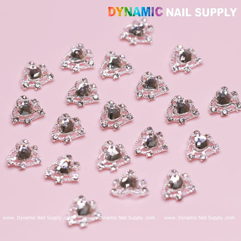 An assortment of elegant, silver heart rhinestone nail charms is displayed in tidy rows against a pastel pink backdrop. Each charm showcases a central crystal heart encircled by elaborate detailing, making them ideal for a beautiful Valentine's nails design. In the top right corner, "Dynamic Nail Supply" is prominently featured in bold letters.
