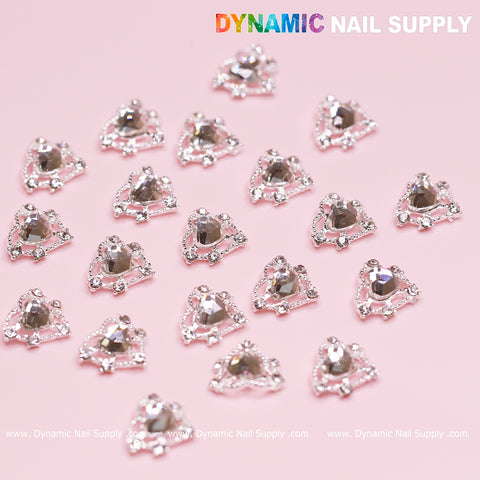 Several silver heart charms featuring crystal rhinestones are arranged on a pink background. These embellishments, ideal for Valentine nails design, include a prominent central rhinestone surrounded by smaller stones for a sparkling effect. The text Dynamic Nail Supply is printed in the top right corner.