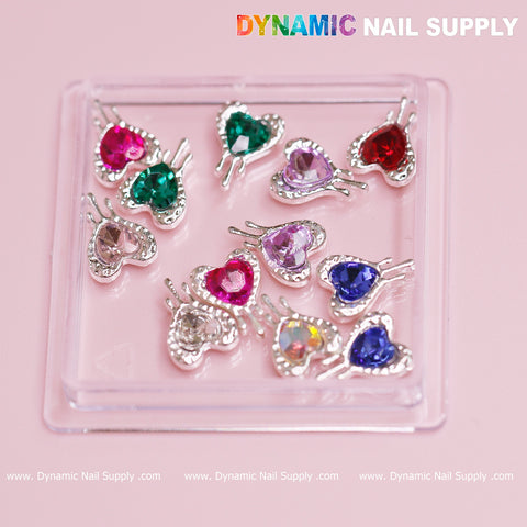 A set of 12 heart-shaped charms with silver edges, adorned with vibrant rhinestones in red, green, pink, blue, and purple hues, beautifully showcased on a clear tray against a soft pink backdrop. Designed for Valentine nail designs, the Dynamic Nail Supply logo is featured prominently at the top in multicolored text.