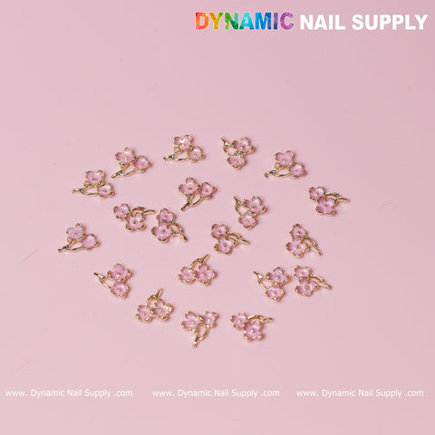 The 20 pcs Pink Cherry Blossom Flower Charms from Dynamic Nail Supply feature delicate small pink flowers with gold accents set against a light pink background. Each blossom, ideal for a romantic Valentine's nail design, boasts five petals and a tiny gem at its center. The brand name "Dynamic Nail Supply" is showcased prominently at the top, with a website link provided below.