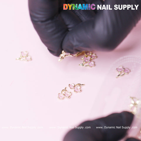 A gloved hand arranges delicate 20 pcs Pink Cherry Blossom Flower Charms for Valentines Nails Design on a light pink surface. Several small flower decorations are scattered, each with gold outlines and tiny blossoms. At the top, the Dynamic Nail Supply logo is featured, repeating at the bottom with their website for Valentines Nails Design inspiration.