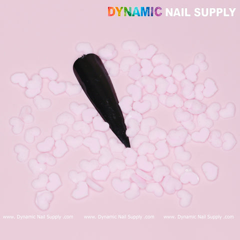 A black pointed nail tip is embellished with a pastel light pink heart charm on a pink background. The brand name, Dynamic Nail Supply, is showcased in colorful letters at the top and repeated in white at the bottom corners, creating an ideal vibe for Valentine nails using the 90 pcs Pastel Light Pink Heart Charm for Valentine Nails Design.