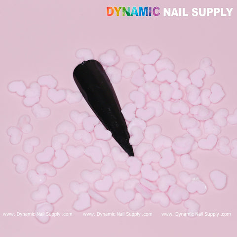 A black false nail is displayed on a soft pink background, embellished with pastel light pink heart charms perfect for Valentine nails. The top right corner showcases the text "Dynamic Nail Supply" in vibrant white lettering.