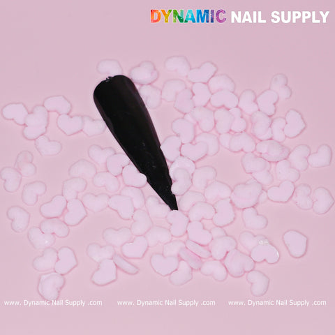 A black nail sample is nestled among small, pastel light pink heart charms scattered on a light pink background, ideal for Valentine Nails. The top right corner showcases "Dynamic Nail Supply" in vibrant letters, while the website URL for Dynamic Nail Supply is elegantly repeated in white text along the bottom.