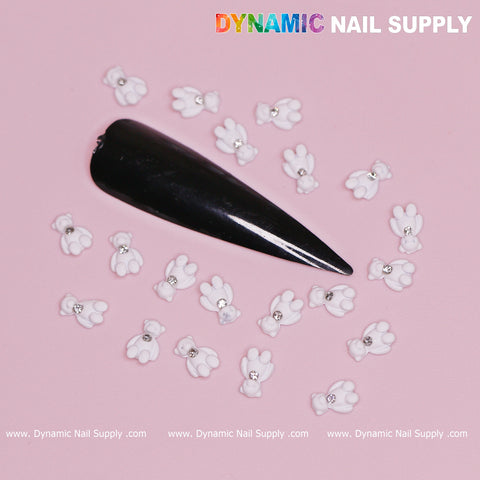 An elongated, glossy black nail with a pointed tip features multiple white teddy bear paw charms, each adorned with a tiny rhinestone. The soft pink background complements the design, complete with the Dynamic Nail Supply logo at the top right—ideal for a Valentine's Day nail look.
