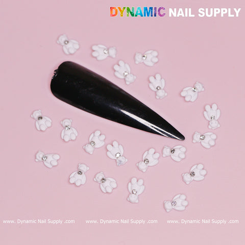 A black pointed false nail is showcased on a pink backdrop, surrounded by small, white 3D paw print nail art adorned with tiny gems. The centerpiece of the design features a charm from the "20 pcs White Teddy Bear Charm for Valentines Nail Design" collection. In the top right corner, "Dynamic Nail Supply" is prominently displayed in colorful and white lettering.