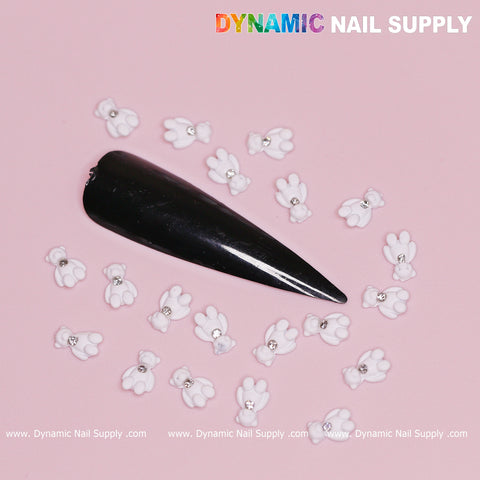 A long, glossy black stiletto nail tip is embellished with small white 3D teddy bear charms, each adorned with a rhinestone. Set against a soft pink background, the design exudes charm reminiscent of a Valentine's nail design. The top right corner showcases "Dynamic Nail Supply" in colorful letters.