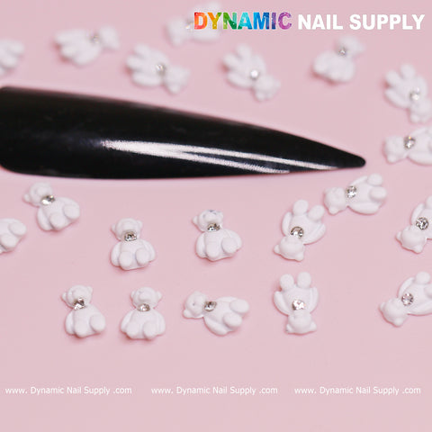 Close-up of Dynamic Nail Supply's 20 pcs White Teddy Bear Charm for Valentine's nail design, featuring rhinestone details, arranged on a pink background. A glossy black nail is partly visible. The branding text "Dynamic Nail Supply" appears at the top, with website URLs repeated along the bottom edge.