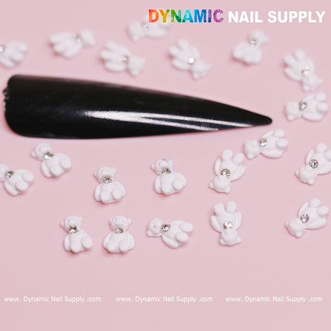 A black, elongated fake nail features small, white 3D bunny-shaped embellishments and silver gem details similar to those in a whimsical Valentine's nail design using the 20 pcs White Teddy Bear Charm for Valentine's Nail Design. The Dynamic Nail Supply brand logo appears in multi-colored letters against a pink backdrop, with their website displayed below.