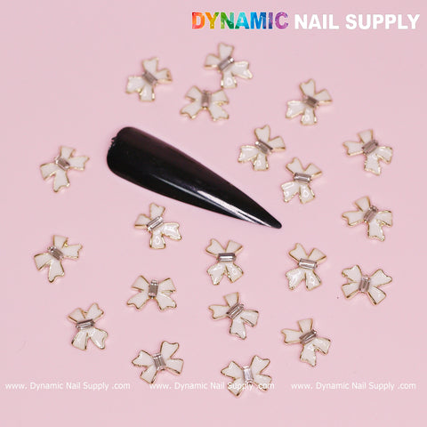 A black pointed false nail embellished with luxury white bow charms detailed with golden contours on a pink background, ideal for chic Valentine's nail designs. "Dynamic Nail Supply" is gracefully displayed along the bottom and top right corner, accompanied by a web address underneath.