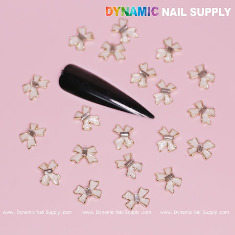 A collection of 20 luxury white bow charms, elegantly trimmed with gold, embellishes a sophisticated black, almond-shaped nail display set against a pink background. At the top, "Dynamic Nail Supply" is prominently featured in vibrant colors, making it perfect for this season's Valentine's nail designs. Website URLs are displayed along the bottom for easy access.