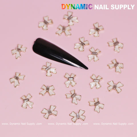 A set of 20 pcs Luxury White Bow Charms with Golden Contours from Dynamic Nail Supply is gracefully displayed on a pink surface. Among the charms, a single black false nail captures attention. The brand name, Dynamic Nail Supply, is prominently positioned at the top and bottom, enhancing the appeal for Valentine's nail designs.