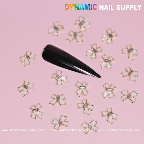 A black, almond-shaped nail tip features an array of gold-tone flower charms and luxurious white bow charms with golden contours against a pink background. The brand name, Dynamic Nail Supply, is elegantly displayed in the top right corner, making it perfect for Valentine's nail designs.