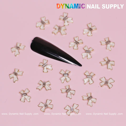 A black artificial nail sits atop a pink surface, featuring 20 luxurious white bow charms with golden contours, perfect for Valentine's nail designs. "Dynamic Nail Supply" is elegantly displayed in the top right corner, while www.dynamicnailsupply.com adorns the bottom, providing inspiration for your nail creations.