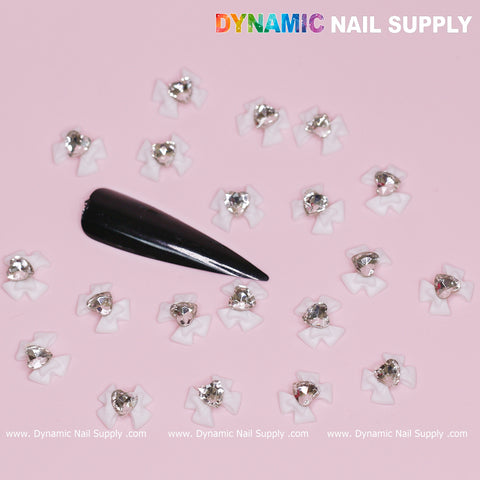 Luxury White Bow Charms engraved with crystal clear hearts are scattered on a pink background alongside white bows adorned with silver rhinestones. An elegant black stiletto nail tip is placed among the charming bows. The brand name "Dynamic Nail Supply" is displayed in colorful letters at the top and repeated below, capturing the essence of enchanting Valentines nail designs.