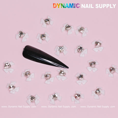 A black stiletto nail displays multiple tiny, white bow-shaped embellishments adorned with crystal-clear heart details on a pink background. The text "Dynamic Nail Supply" is elegantly positioned at the top in a vibrant gradient. Elevate your style with Valentines Nail Designs and explore more at www.DynamicNailSupply.com below.