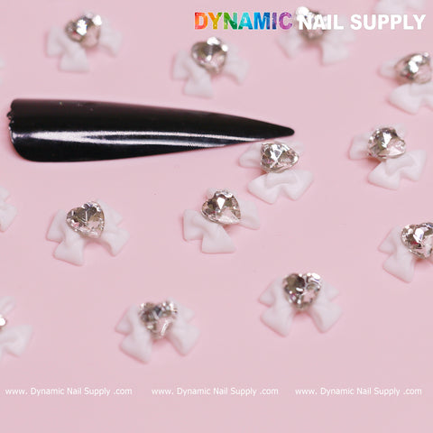 A set of 20 luxury white bow charms engraved with crystal clear heart jewels is artfully arranged on a pink background. Amid the decorations, a black nail tip stands out, showcasing elegant Valentine's nail designs. The brand name Dynamic Nail Supply is prominently displayed at both the top and bottom of the image.
