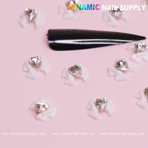 A pink surface showcases luxurious white bow charms adorned with crystal clear heart-shaped rhinestones, elegantly arranged around a long, glossy black nail tip. The image features a watermark of Dynamic Nail Supply, with colorful lettering at the top and white text at the bottom.