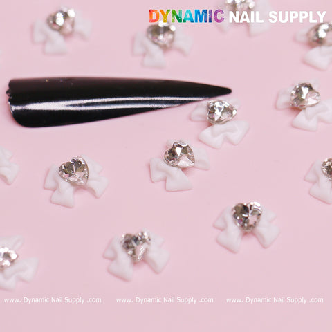 A black nail sample rests on a pink surface, embellished with luxury white bow charms engraved with a crystal clear heart, making them ideal for Valentine's nail designs. The top right corner prominently features "Dynamic Nail Supply" in vibrant letters, while the website URL is displayed at the bottom.