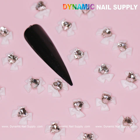 Close-up of a pink surface featuring a black stiletto nail tip adorned with Luxury White Bow Charms engraved with Crystal Clear Heart accents, perfect for Valentine's Nail Designs, accompanied by sparkling silver gemstones. The brand name, Dynamic Nail Supply, is printed across the top and bottom of the image.