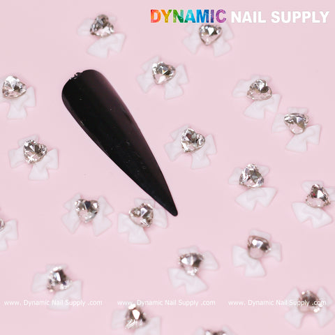 A black coffin-shaped nail tip is decorated with luxury white bow charms, engraved with Crystal Clear Heart accents from Dynamic Nail Supply, and silver rhinestones, creating a stunning contrast against a pink background. The elegance is further enhanced by the placement of Dynamic Nail Supply's brand name on the top right and bottom of the image.