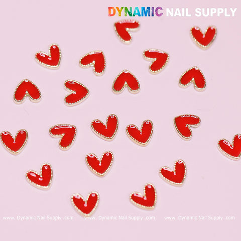 Small red heart-shaped nail art charms with golden borders are scattered on a pink surface. In the upper right corner, the brand name Dynamic Nail Supply is displayed, while the bottom edge features their website repeatedly: www.DynamicNailSupply.com—a perfect addition for Valentine nails design using their 20 pcs Heart Charm with Golden Borders.