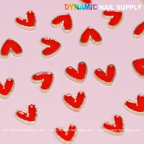 The image features small, red, heart-shaped nail art charms with golden borders, scattered across a light pink background. In the top right corner, bold and colorful letters display "Dynamic Nail Supply," while the website URL www.DynamicNailSupply.com is positioned at the bottom edge. Ideal for a Valentine nails design using the 20 pcs Heart Charm with Golden Borders by Dynamic Nail Supply!