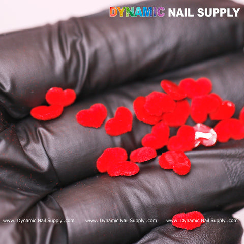 A hand clad in black gloves holds several charms from Dynamic Nail Supply, specifically the "90 pcs Pure Red Resin Heart Shape Charm for Valentine Nails Design." The background features the vibrant text "DYNAMIC NAIL SUPPLY," with the website subtly displayed below as www.DynamicNailSupply.com.
