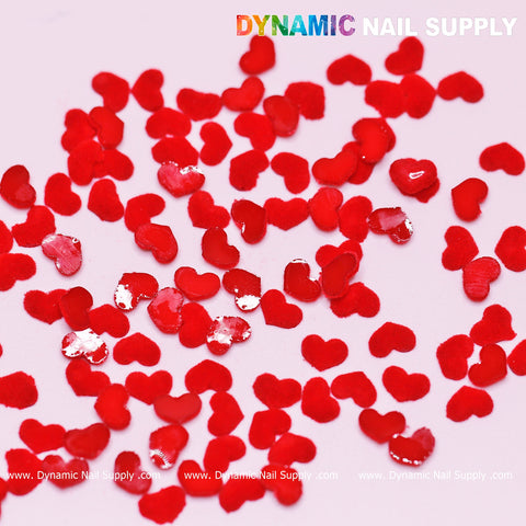 A light pink surface is beautifully adorned with Pure Red Resin Heart Shape Charms from the 90-piece set, making it ideal for crafting romantic Valentine Nail Designs. These heart-shaped charms shine with a glossy finish. Displayed prominently at the top is the logo of Dynamic Nail Supply in vibrant colors, while the brand's website URL appears subtly at the bottom in small white text.