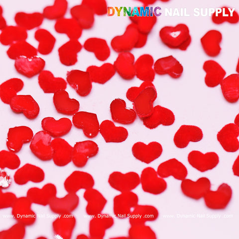 A close-up shot features small, vibrant red heart-shaped nail art decorations from Dynamic Nail Supply, ideal for a Valentine nails design, scattered on a white surface. The hearts are part of the 90 pcs Pure Red Resin Heart Shape Charm set and vary slightly in size and orientation. At the top, the brand name is highlighted in colorful, bold letters.