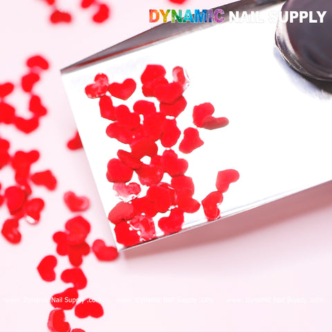 Close-up of metallic tweezers holding a tiny heart-shaped charm from the 90 pcs Pure Red Resin Heart Shape Charm set, designed for Valentine Nails Design. More hearts are scattered on a white surface, creating a perfect backdrop for your nail art. The top right corner showcases the Dynamic Nail Supply brand name in gradient colors.