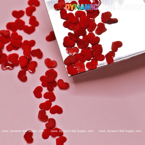 Red heart-shaped confetti cascades from a metallic surface onto a light pink background, capturing the essence of Valentine Nails Design. The top right showcases the text Dynamic Nail Supply in vibrant letters. For more information about the 90 pcs Pure Red Resin Heart Shape Charm for Valentine Nails Design, visit www.DynamicNailSupply.com.