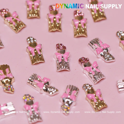 A close-up view of decorative nail charms highlights the 20 pcs Lipstick Charms with Pink Bows for Valentine's Nail Art Design, artfully arranged on a pink surface. Each charm resembles a bottle adorned with a pink bow and a rhinestone cap, shimmering in gold and silver. The brand name "Dynamic Nail Supply" stands out in vibrant letters, making these charms perfect for inspiring Valentine's nail art designs.