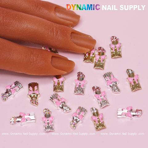 A manicured hand hovers near scattered lipstick charms on a light pink surface. These charming adornments, ideal for Valentine's nail art, showcase small pink bows atop silver and gold bases adorned with crystals. The brand name "Dynamic Nail Supply" is elegantly printed at the top of the image.