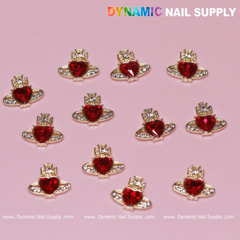Twelve Vivienne Westwood Vintage Red Heart Charms from Dynamic Nail Supply rest on a pink surface. Each charm is adorned with a radiant heart gem in the center, surrounded by sparkling clear rhinestones. The background subtly displays the Dynamic Nail Supply logo in the top right corner and along the bottom.