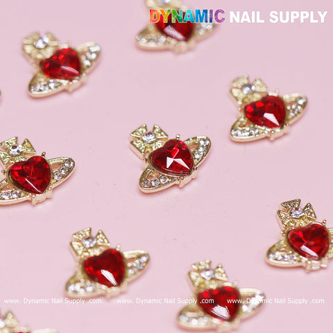 Twelve pieces of Vivienne Westwood Vintage Red Heart Charms for nail art, crafted in the shape of spaceships encapsulating Vivienne Westwood's whimsical style, exhibit a vintage red heart jewel at their core. They are delicately bordered with small clear gemstones and artfully arranged on a pink surface. The top of the background proudly displays the brand name: Dynamic Nail Supply.