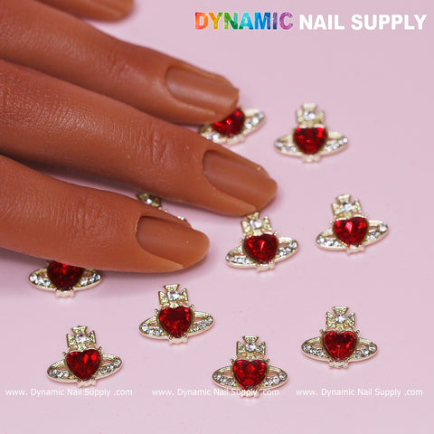 A hand with brown nails rests on a pink surface, adorned with 12 pcs Vivienne Westwood Vintage Red Heart Charms for Nail art designer. These charms feature vintage red heart gems accented with silver orb-like designs and small clear gems that captivate the eye. In the upper-left corner, colorful text proudly showcases Dynamic Nail Supply.