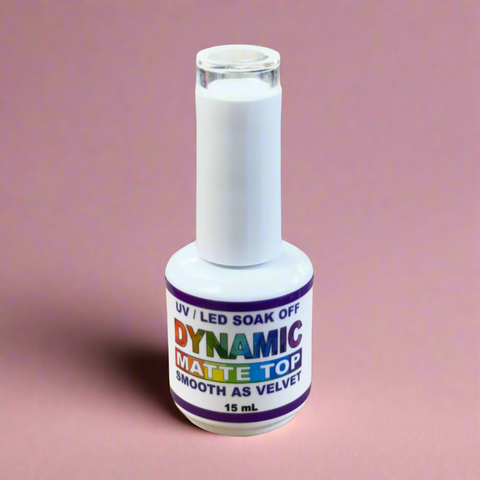 A bottle of Dynamic Nail Supply's Dynamic No-Wipe Matte Top Coat, promising a long-lasting velvet finish, rests against a pink backdrop. The label highlights UV LED Soak Off features and the 15 mL size is clearly visible. A white cap harmonizes with the overall color scheme, while shadows cast leftward add depth and intrigue.